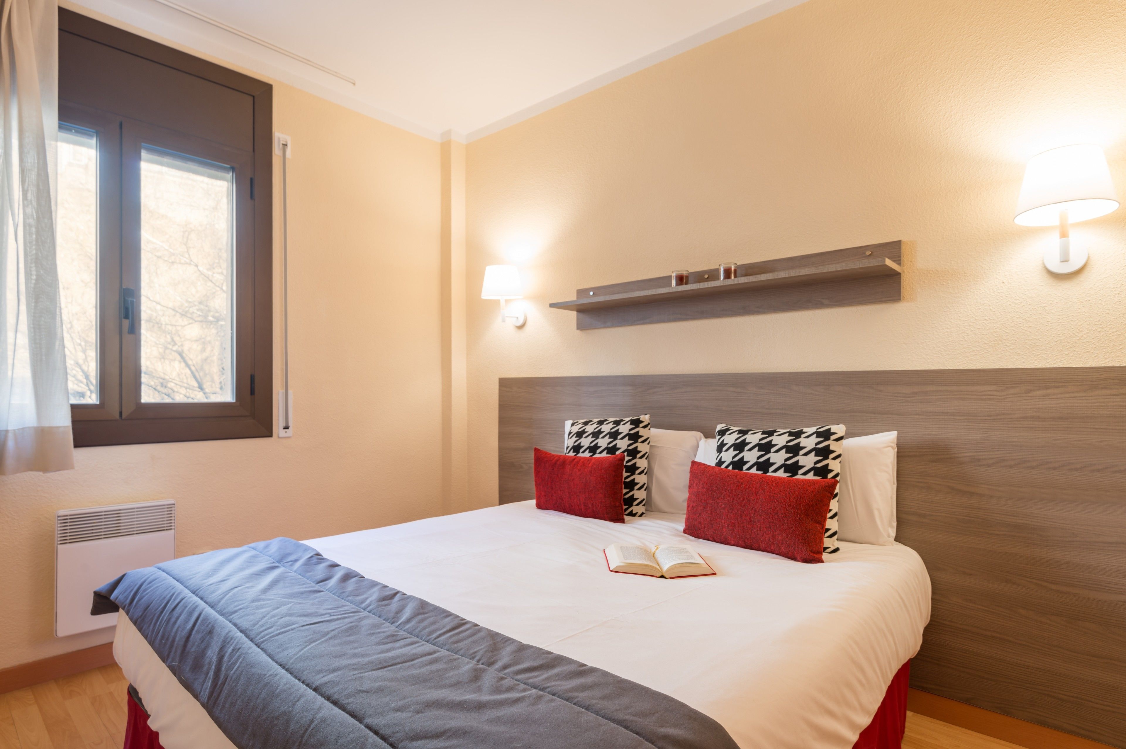 Een netjes opgemaakt bed met een rood-wit kussen, een nachtkastje en een raam met gordijnen.