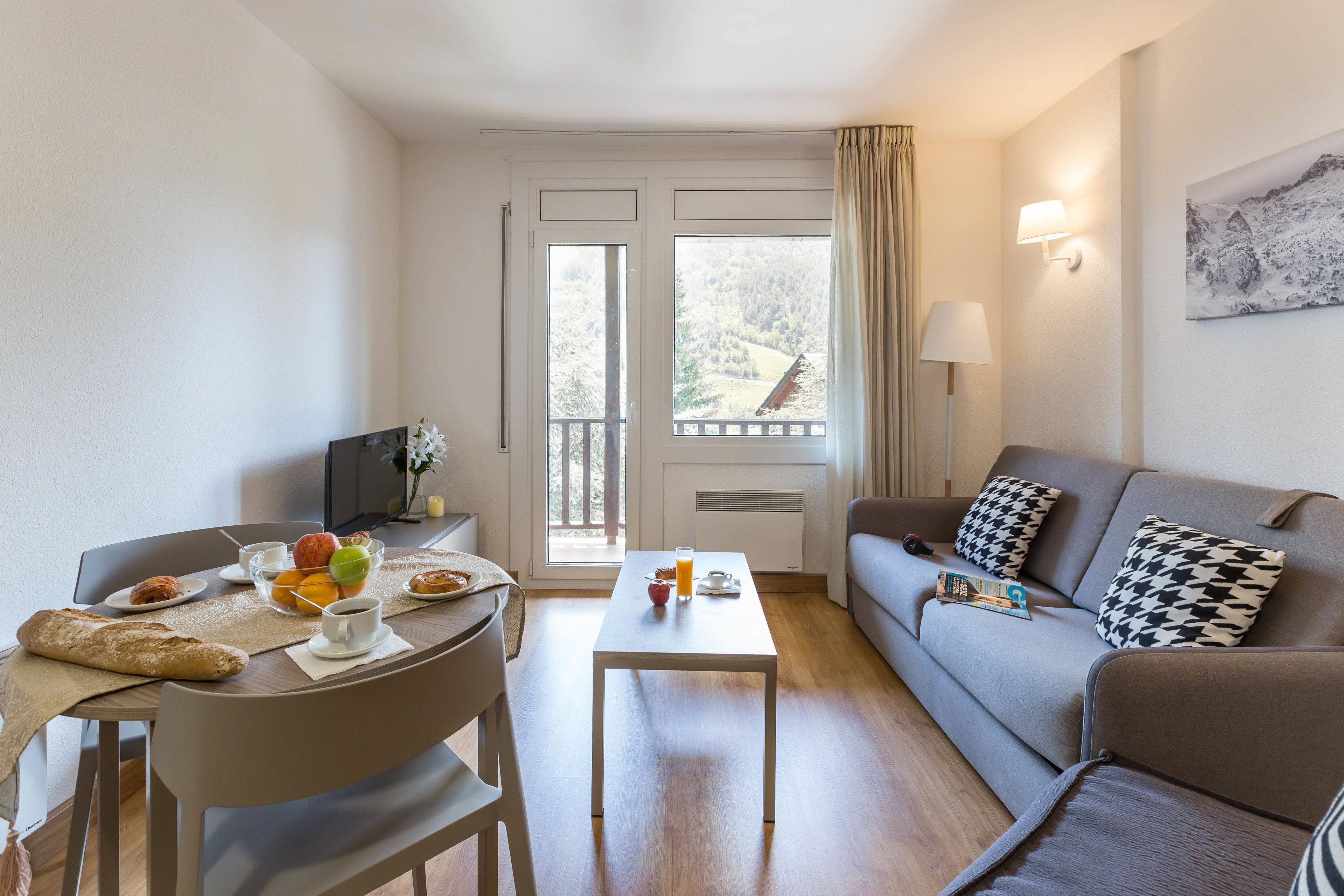 A modern living room with a dining area, a sofa, a table with breakfast items, and a view of a mountainous landscape through the windows.