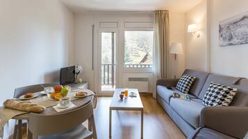 A modern living room with a dining area, a sofa, a table with breakfast items, and a view of a mountainous landscape through the windows.