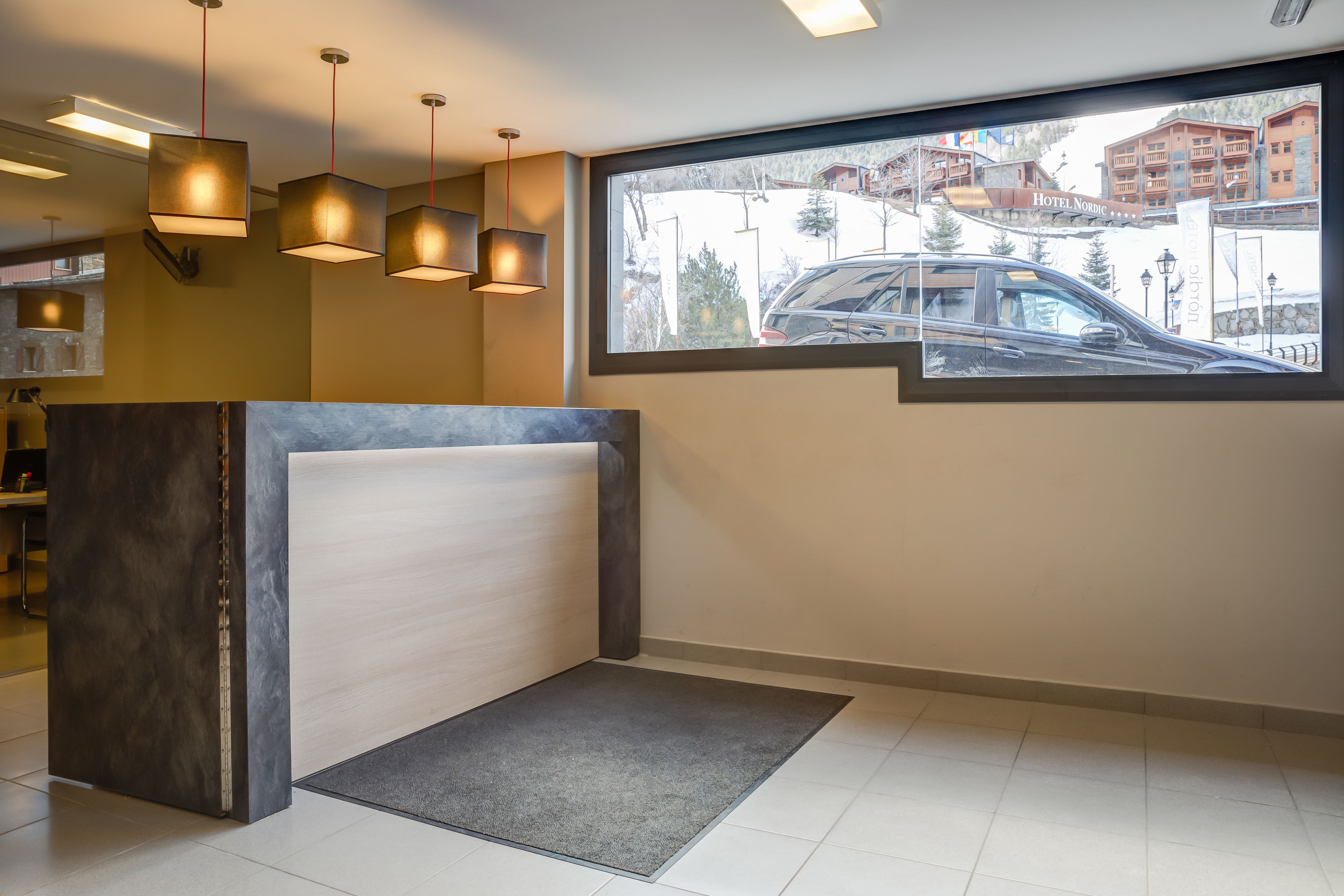 Zona de recepción de un hotel con diseño moderno, que incluye un mostrador de recepción, varias lámparas colgantes y un amplio ventanal con vistas al hotel y sus alrededores.
