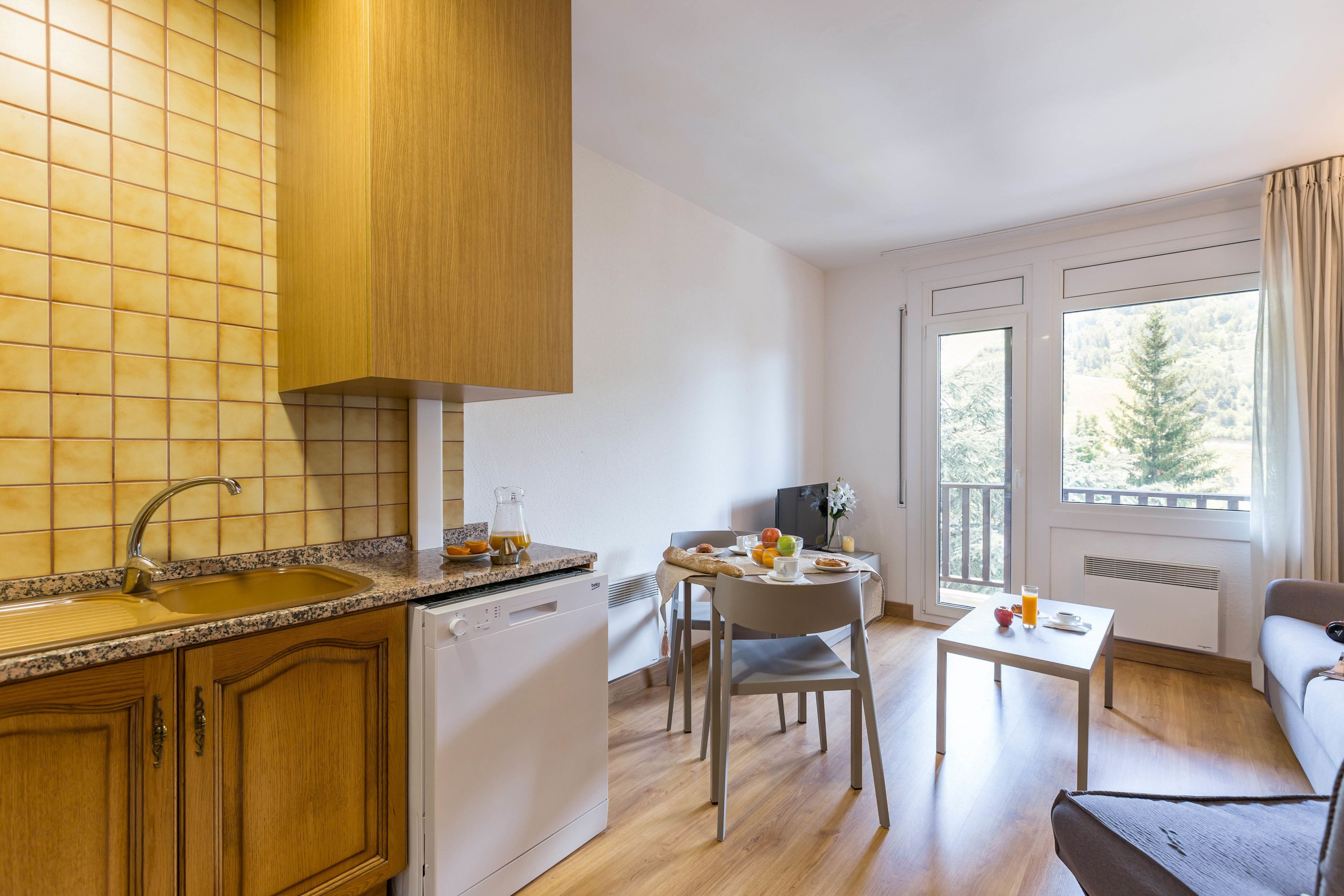 Cocina moderna con encimera de granito, armarios de madera y lavavajillas. Cuenta con una zona de comedor con mesa, sillas y sofá. La habitación tiene ventanas amplias con vistas a árboles.