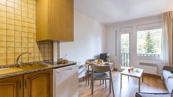 Cocina moderna con encimera de granito, armarios de madera y lavavajillas. Cuenta con una zona de comedor con mesa, sillas y sofá. La habitación tiene ventanas amplias con vistas a árboles.