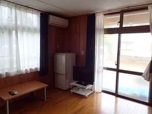 A small room with a wooden bench, a white refrigerator, a TV on a mobile stand, and a sliding glass door to the outside.