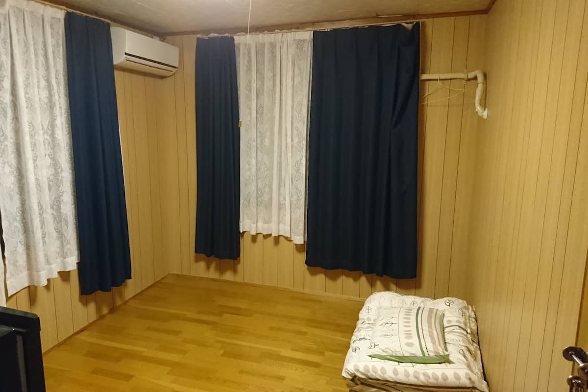 A room with wooden panel walls, a wooden floor, a bed with a white and green patterned blanket, and two windows with blue and white curtains.