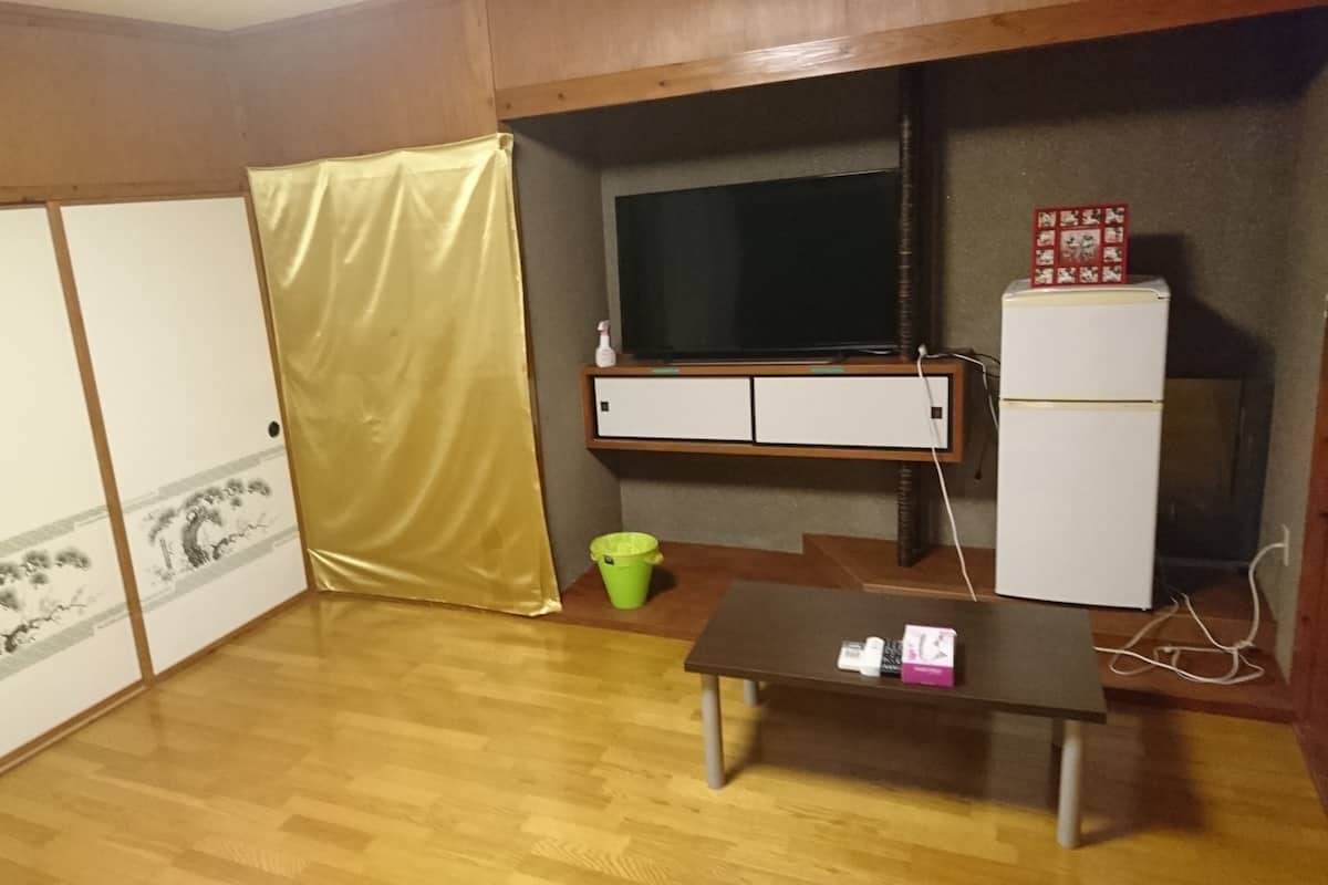 A room with a wooden floor, a television on a shelf, a small table, a green trash bin, and a white refrigerator.