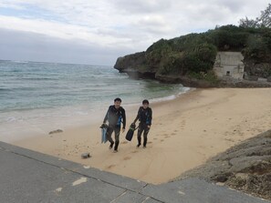 Two people walking on a sandy beach with a rocky cliff and ocean in the background. - Sea Blue (Onna)