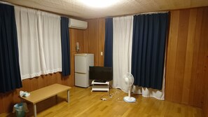 A room with wooden walls, a white air conditioner, a white refrigerator, a TV on a stand, a fan, a wooden bench, and a trash bin. - Sea Blue (Onna)