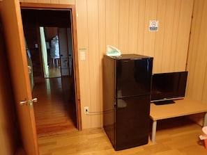 A compact room with a wooden-paneled wall, a black refrigerator, a television on a stand, and a door leading to another room.