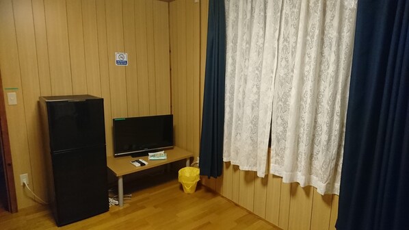 A room with a wooden paneled wall, a black refrigerator, a flat-screen TV on a wooden stand, a trash bin, and a window with white lace curtains.
