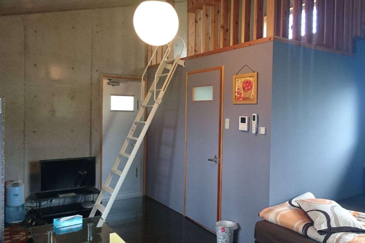 A room with a TV, a ladder, a door, a wall-mounted thermostat, and a window.
