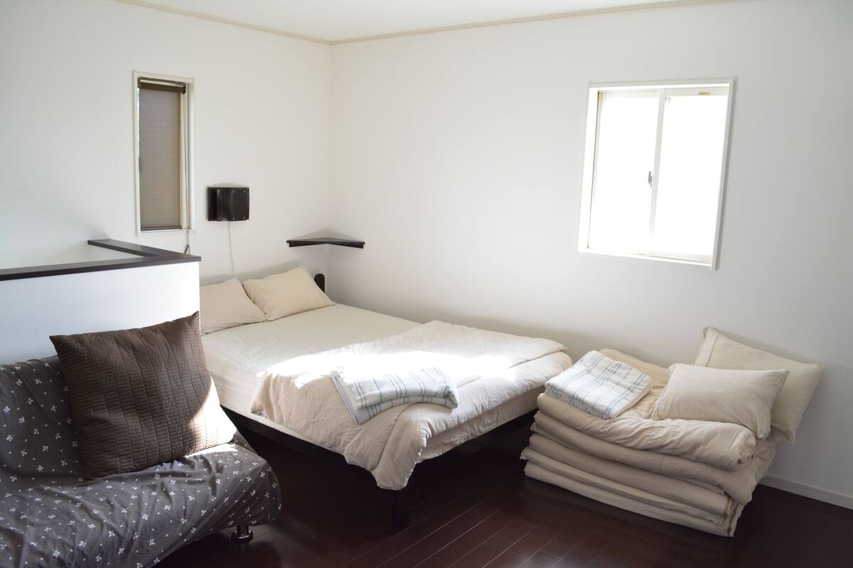 A bedroom with a bed, a bedside table, a chair, a window, and a wall-mounted speaker.