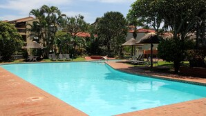 A swimming pool with lounge chairs and umbrellas, surrounded by greenery and buildings.