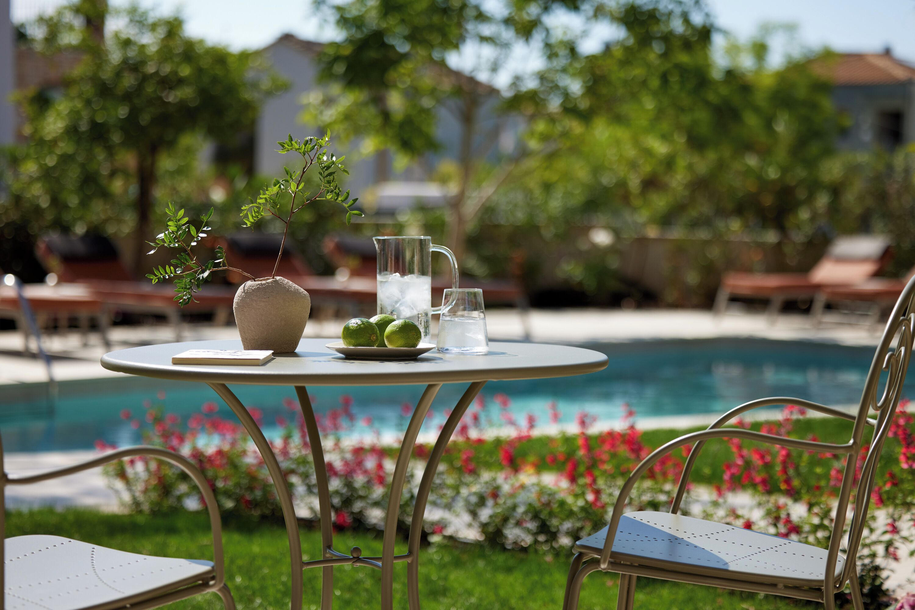 Une table ronde avec une carafe, des verres et une assiette de citrons verts, placée au bord d’une piscine, entourée de chaises longues et de verdure.