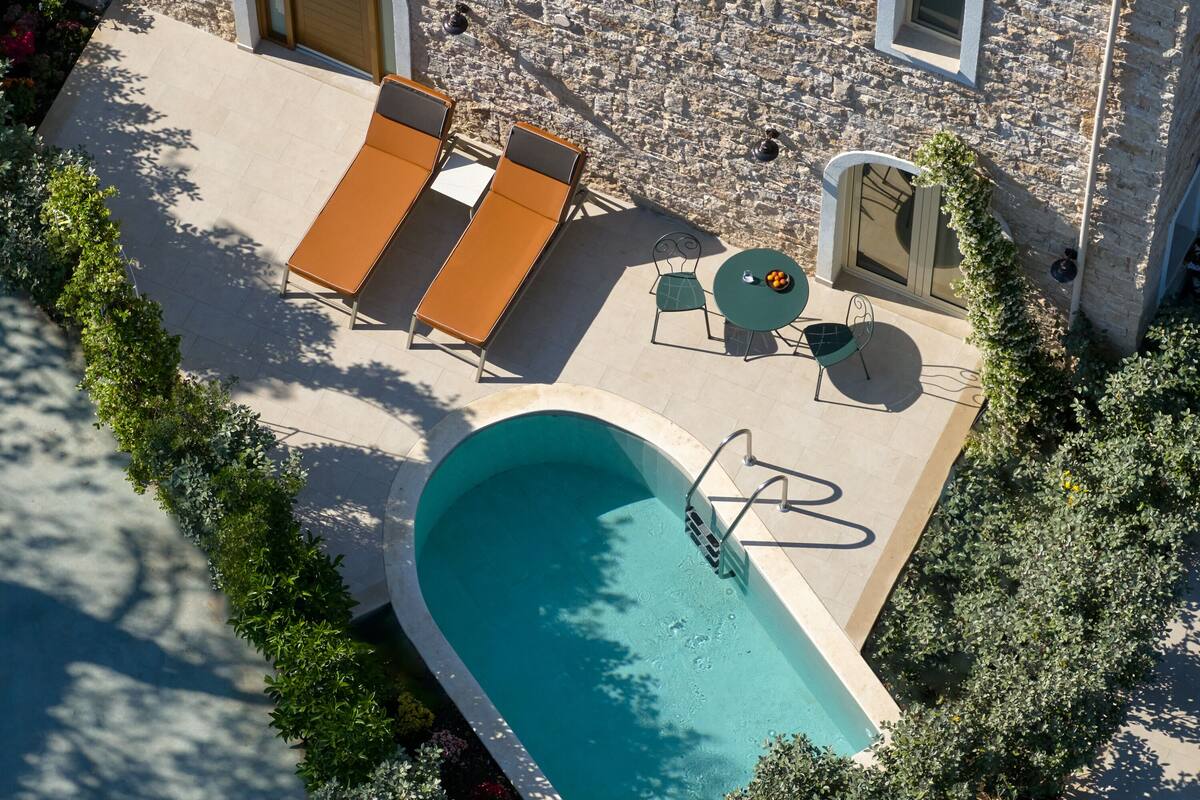 An outdoor swimming pool area with lounge chairs and a table, surrounded by stone walls and greenery.