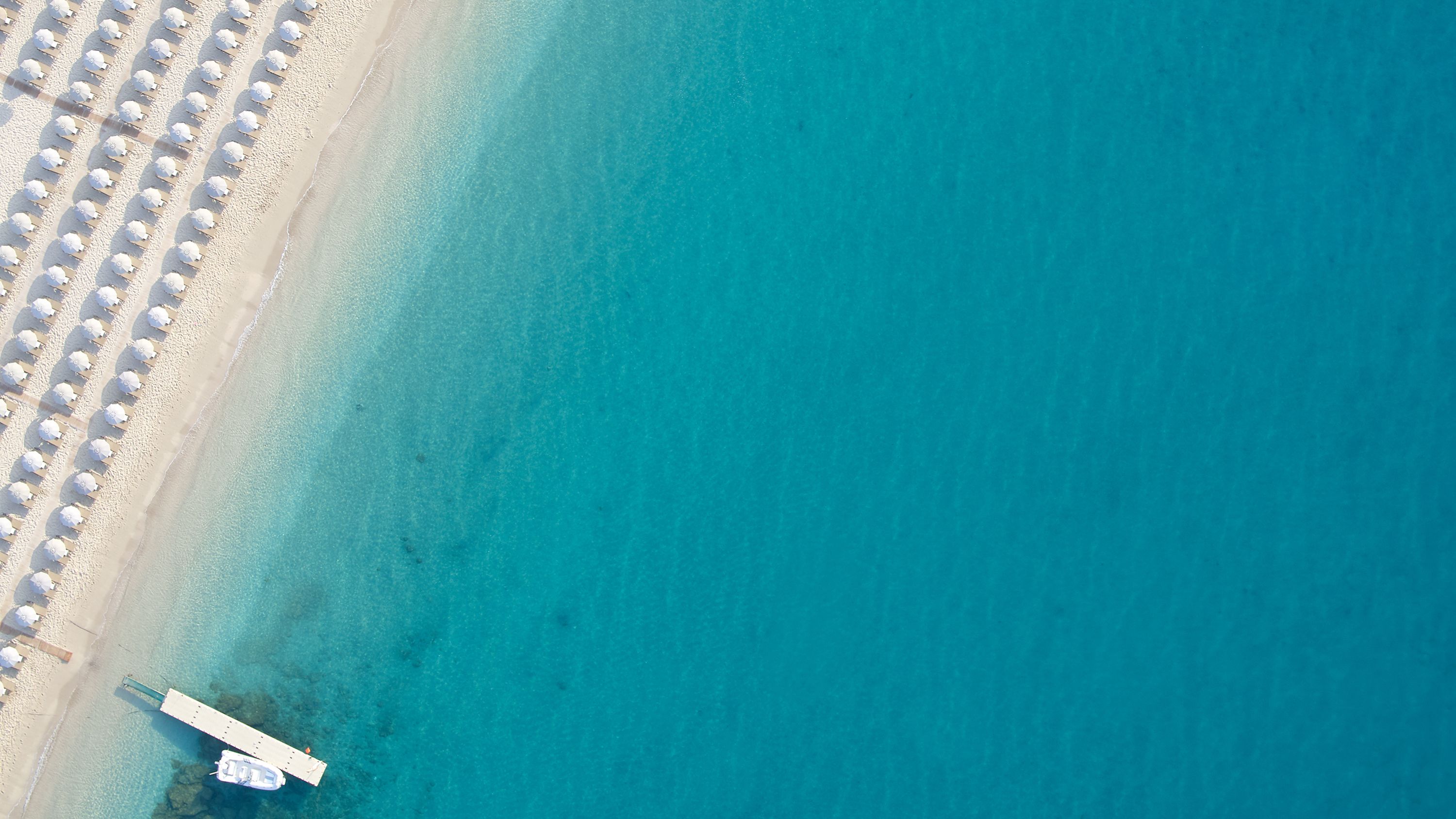 An aerial view of a beach with rows of white umbrellas and a pier extending into the turquoise water.