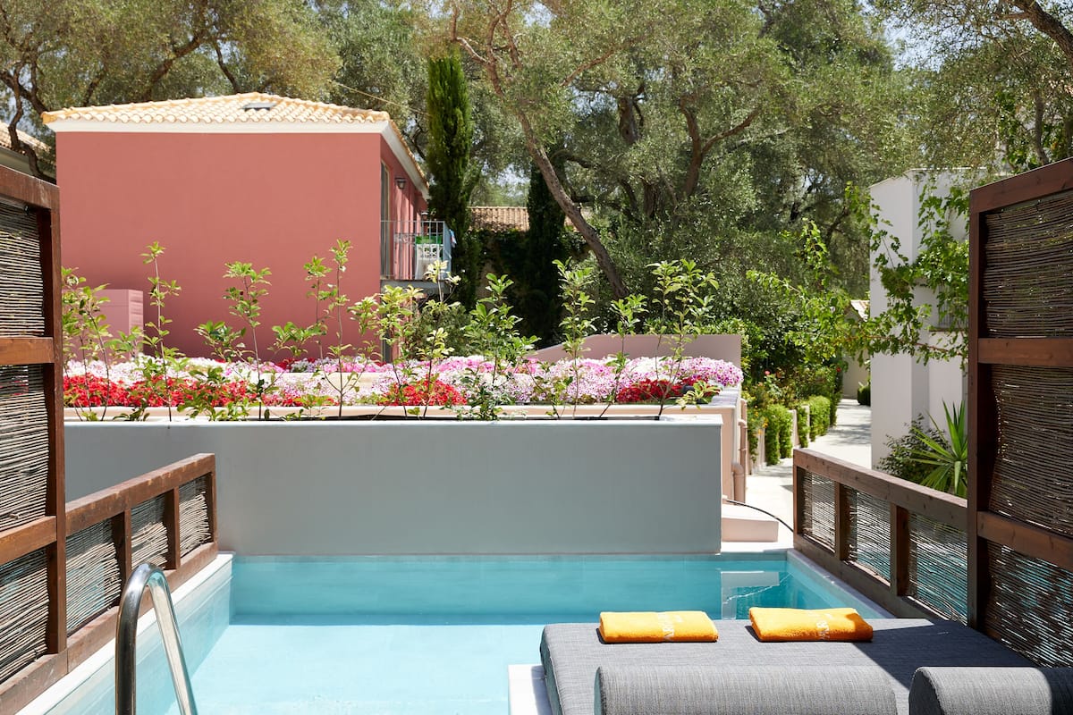 A pool area with two grey lounge chairs, a wooden fence, and a building with a red facade in the background.
