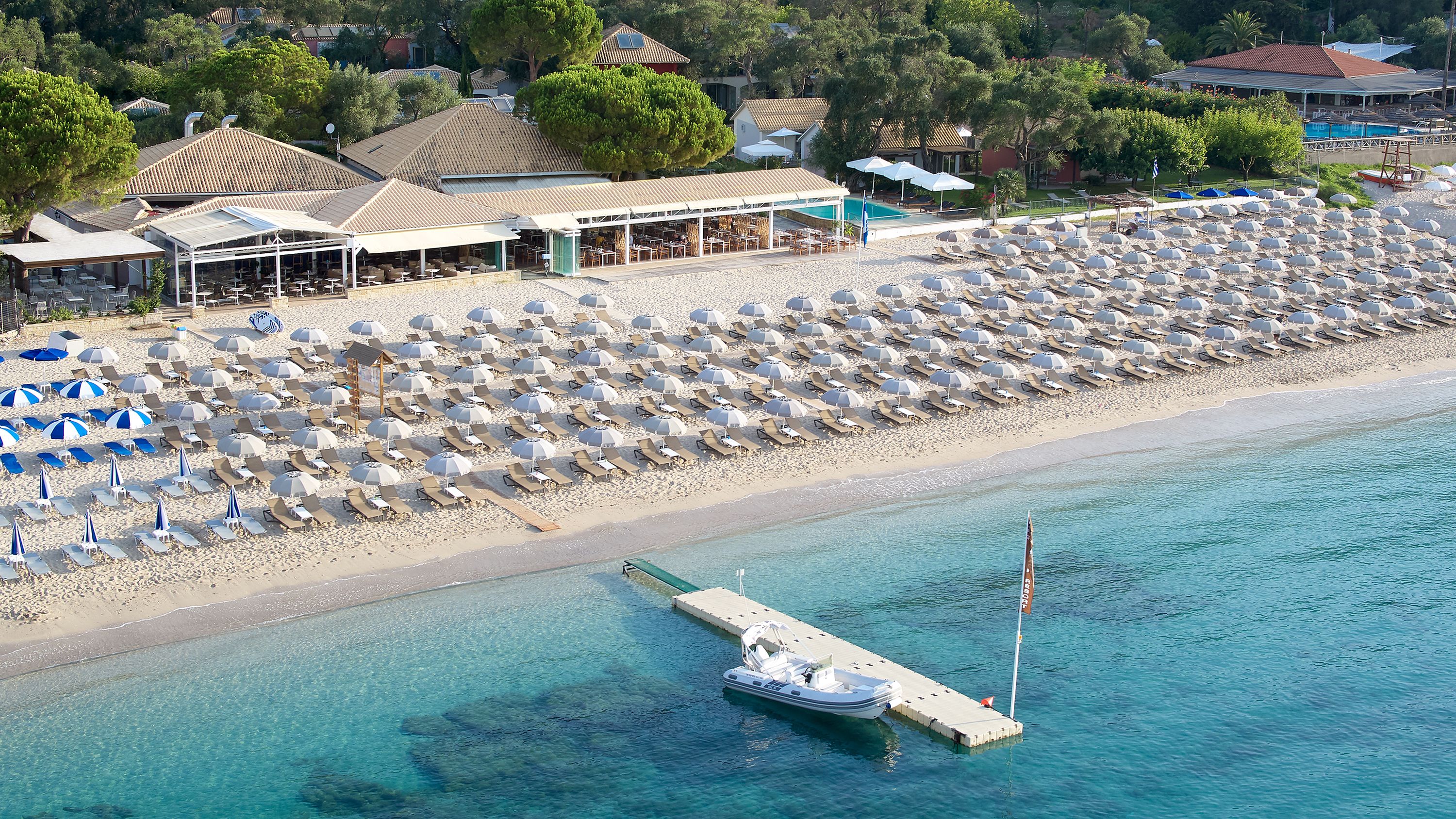 An aerial view of a beach with rows of sun loungers and umbrellas, a dock with a boat, and buildings in the background.