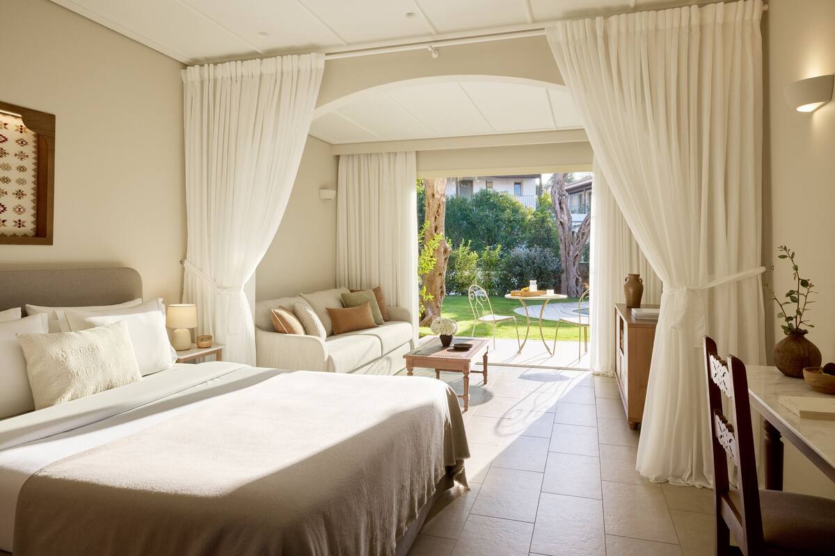 A hotel room with a large bed, a sofa, a small table, and a view of a garden and buildings outside.