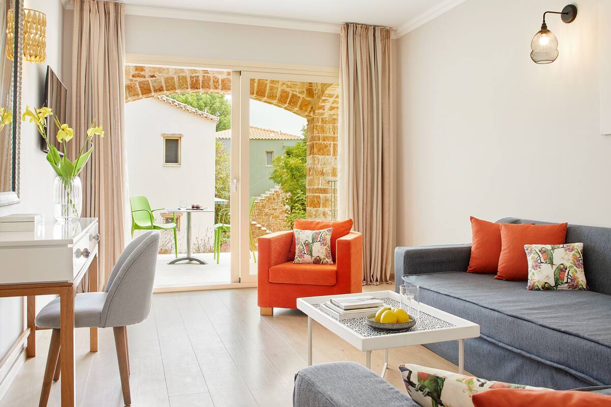 A modern living room with a sofa, armchairs, and a coffee table. There is a dining area with a table and chairs, and a view of a garden through an open door.