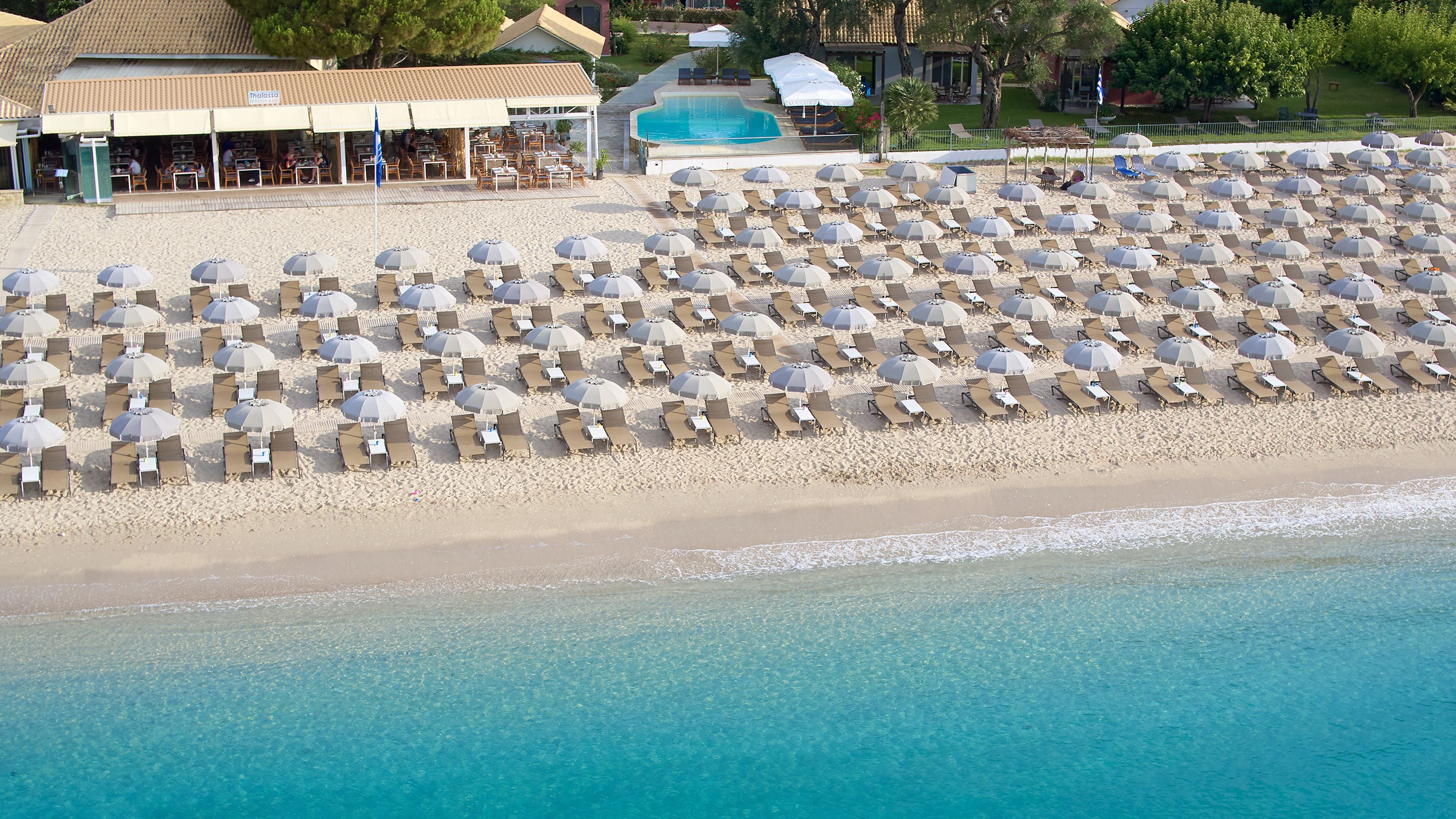 Vue aérienne d’une plage avec de nombreux transats et parasols, une piscine et un restaurant appelé « Thalassa ».