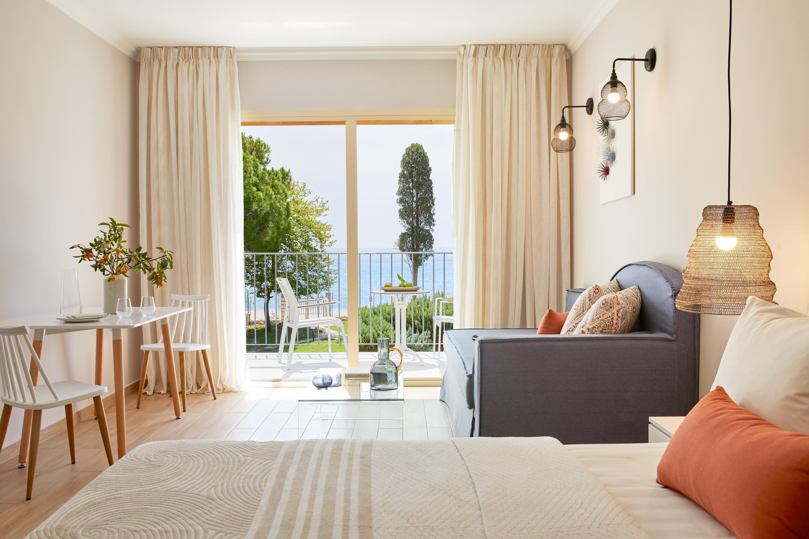 A hotel room with a bed, a sofa, a dining area with chairs and a table, and a balcony with a view of the sea.