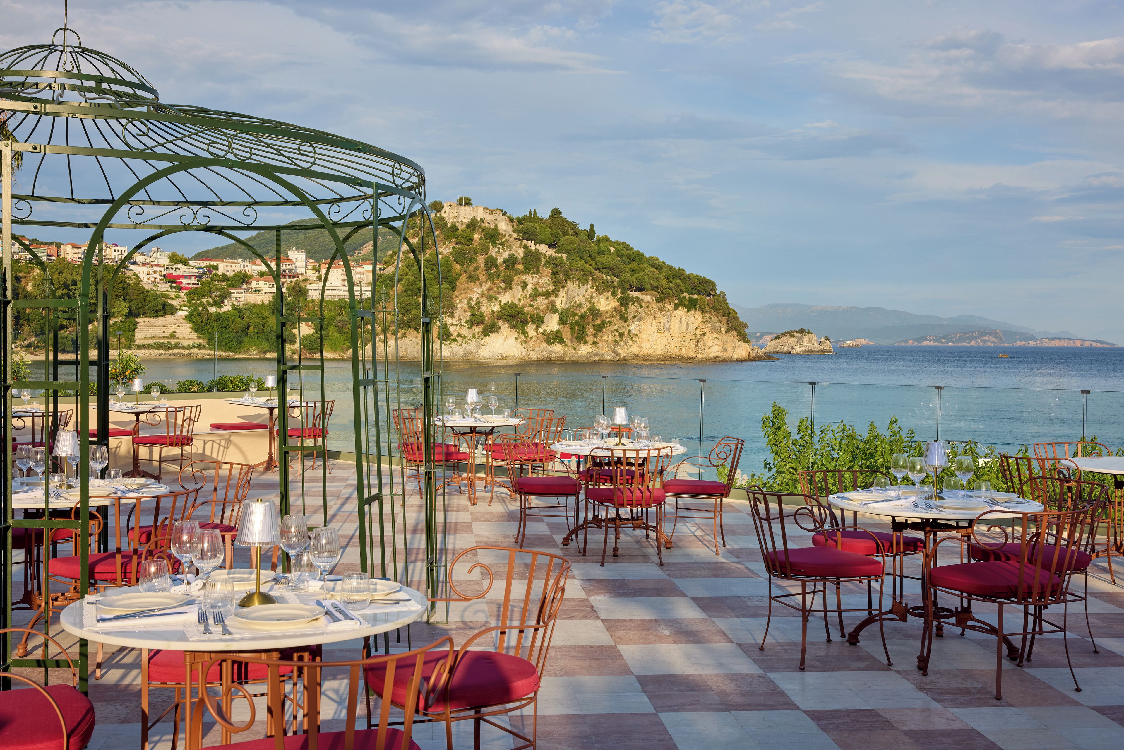 An outdoor dining area with tables and chairs, overlooking a body of water and a hilly landscape.