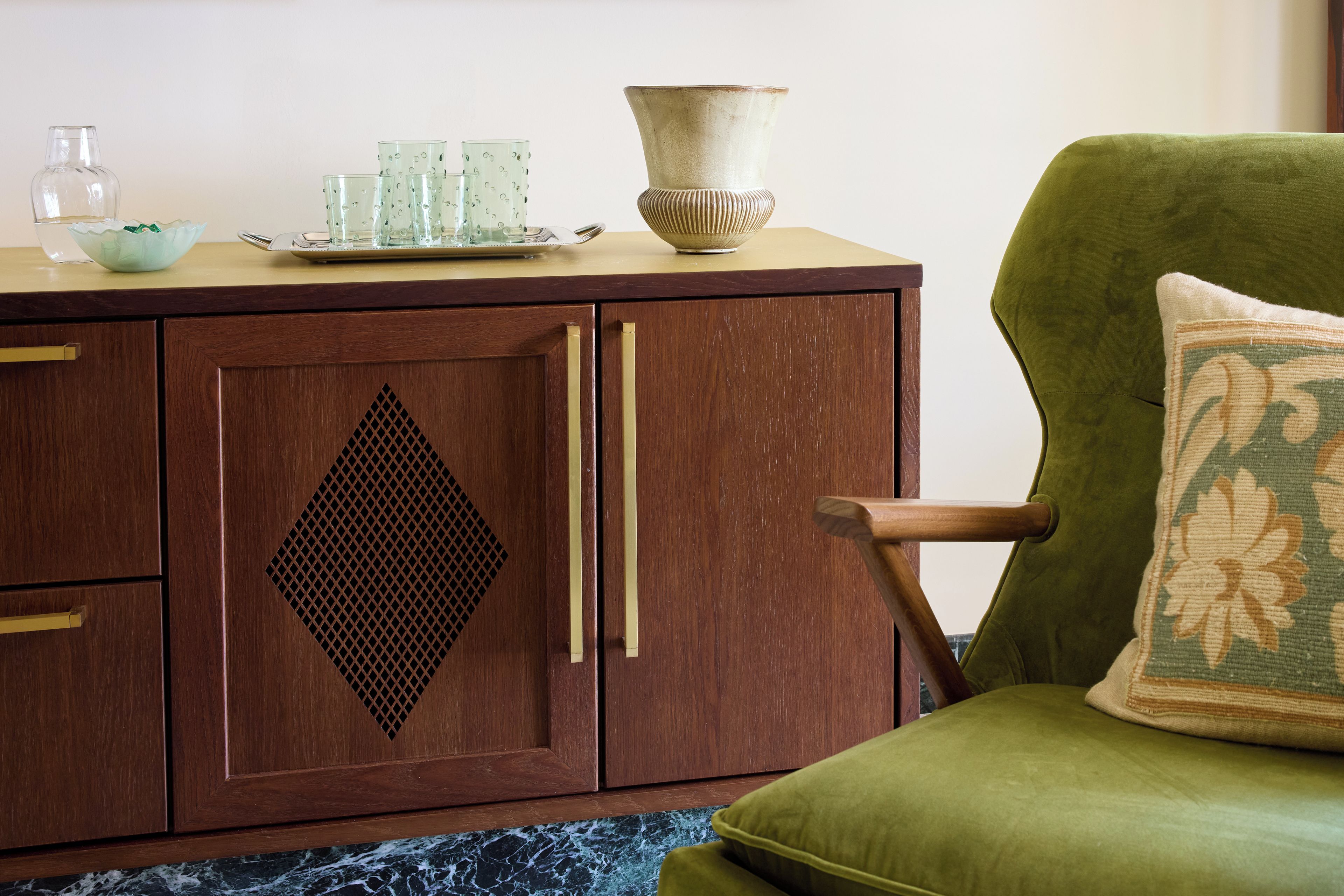 A wooden cabinet with a diamond-shaped cutout, a green armchair with a patterned pillow, and a tray with glassware on top.