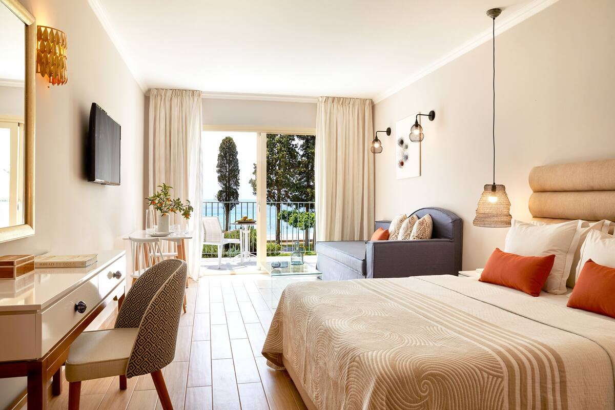 A hotel room with a bed, a desk, a chair, a sofa, a TV, and a balcony with a view of trees and the sky.