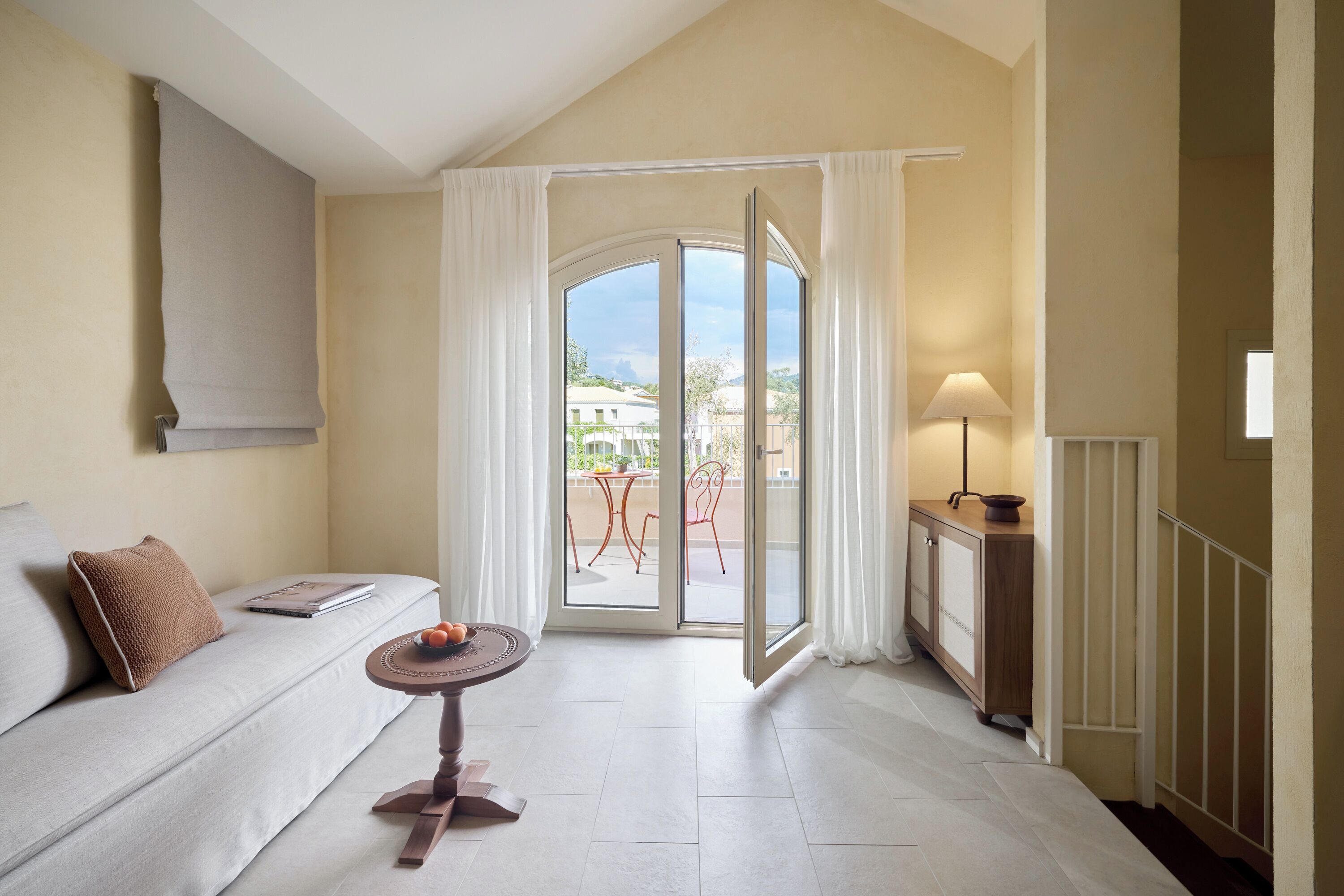 Habitación de hotel con sofá, una mesita con fruta, una lámpara y un ventanal corredero que da a un espacio exterior con mesa y sillas.