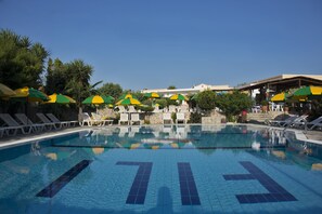 A swimming pool with lounge chairs and umbrellas, surrounded by trees and a building in the background.