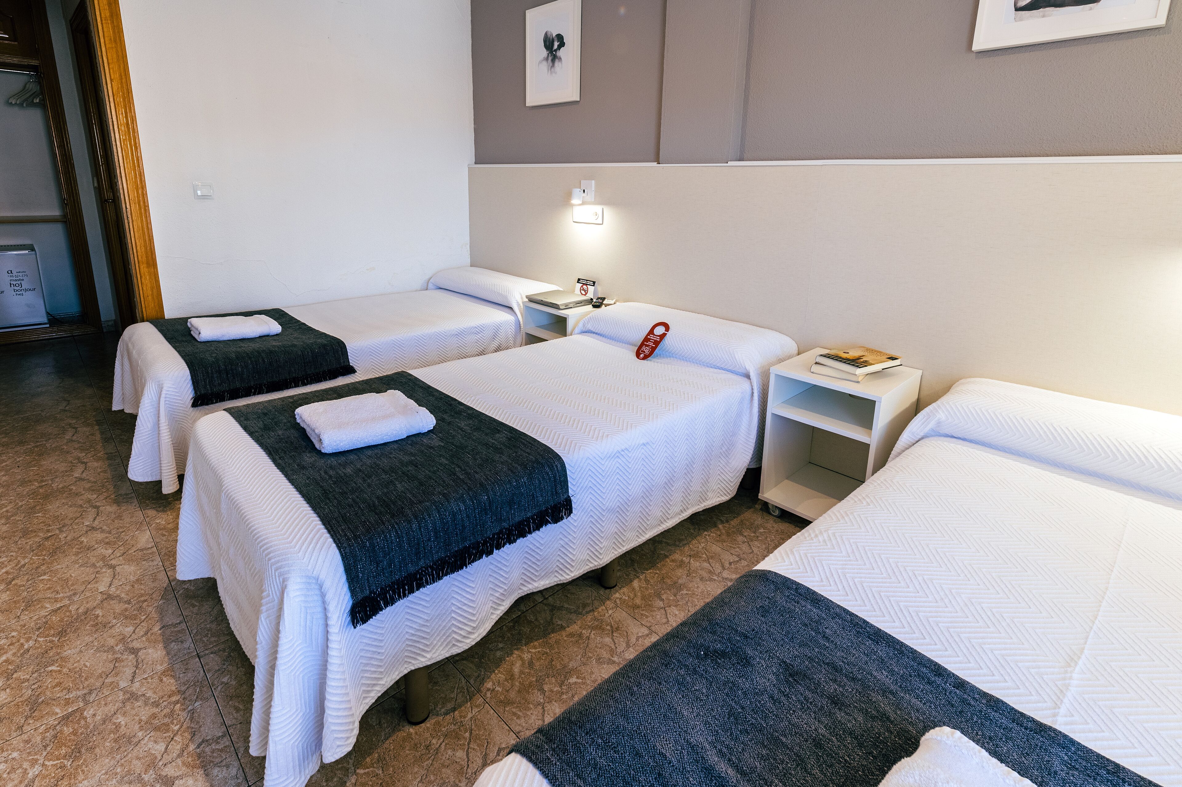 A hotel room with three single beds, each with a white bedspread and a dark blue blanket. There is a nightstand with a lamp and a phone on one side, and a small shelf with books and a framed picture on the other.
