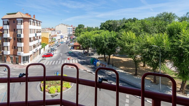 A street view from a balcony with a red railing, featuring a roundabout, parked cars, and buildings with balconies. - Hostal Gran Vía (Pinto)