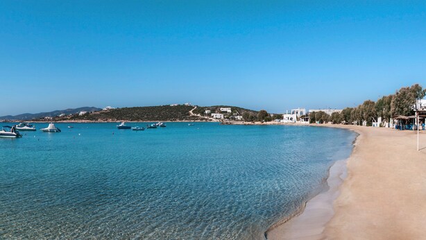 Une plage côtière aux eaux bleues et limpides, avec des bateaux et des bâtiments en arrière-plan.