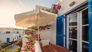 Une table et des chaises en bois, placées sous un parasol blanc sur un balcon orné de fleurs roses et doté d’une porte bleue.