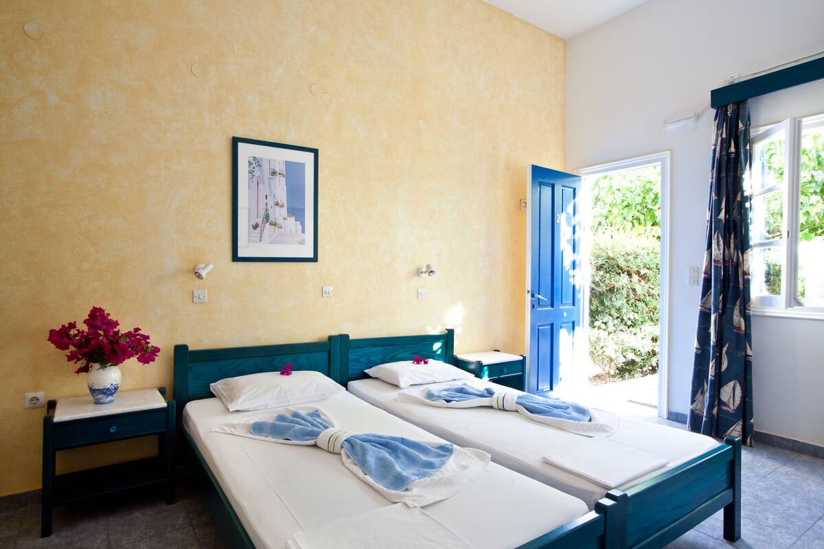 A room with two beds, a vase of flowers, a framed picture on the wall, and a window with curtains.