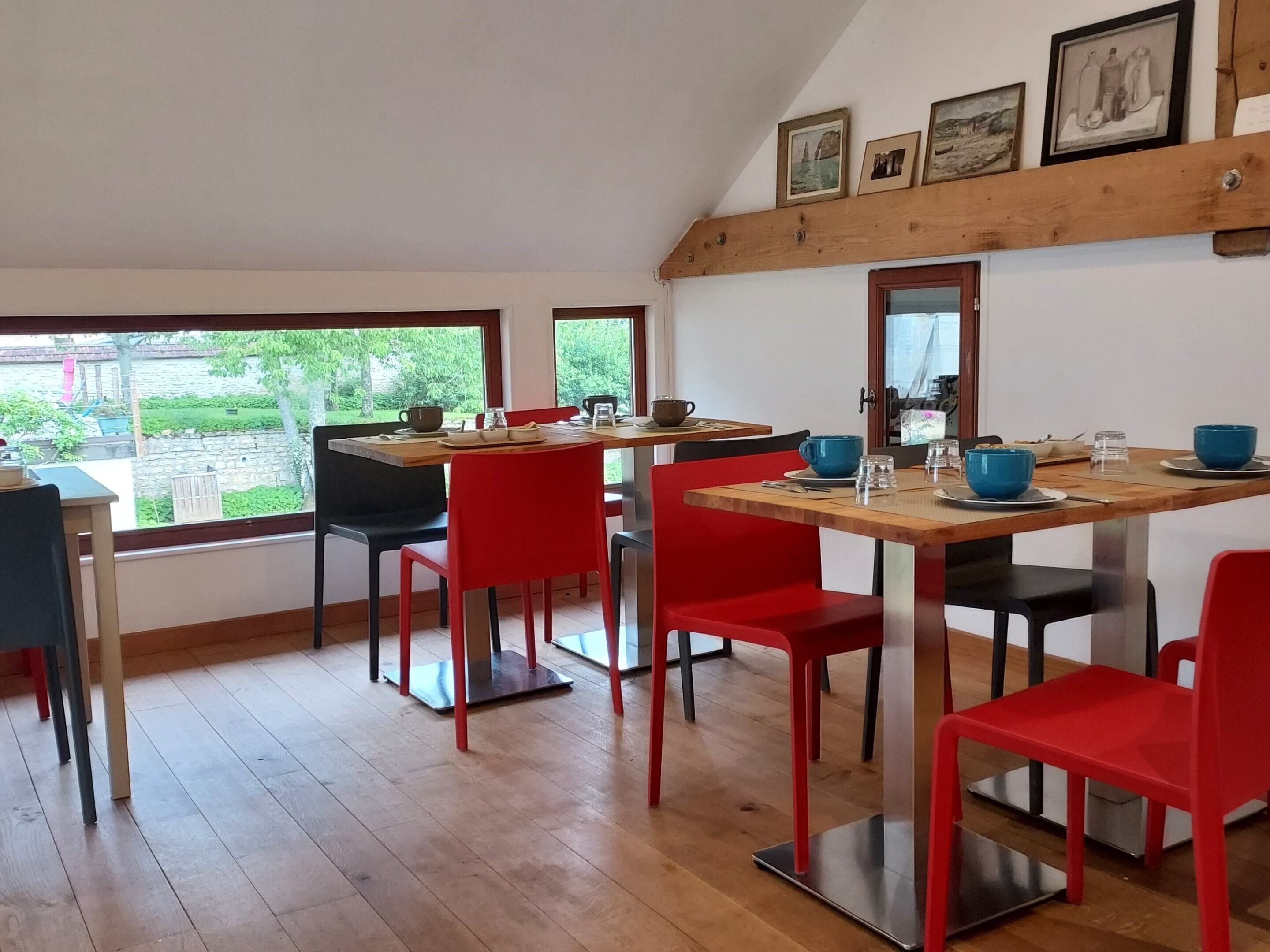 Een eetruimte met houten tafels en rode stoelen, grote ramen met uitzicht op groen en aan de muur hangende foto's.