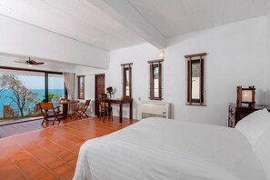 A bedroom with a large bed, a desk with chairs, a view of the sea, and a ceiling fan.