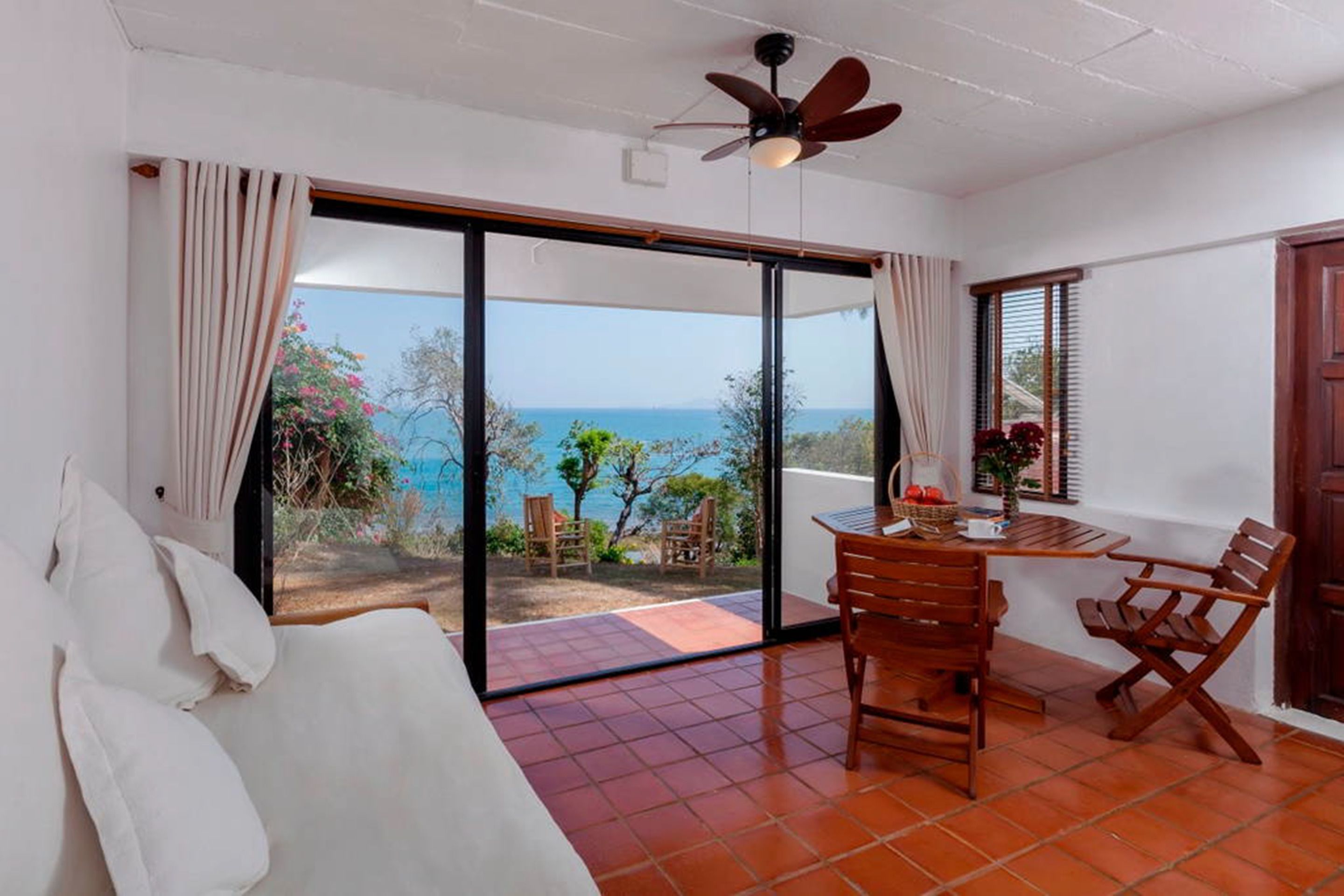 A room with a large sliding glass door leading to a balcony with a view of the sea. There is a ceiling fan, a table with chairs, and a sofa.