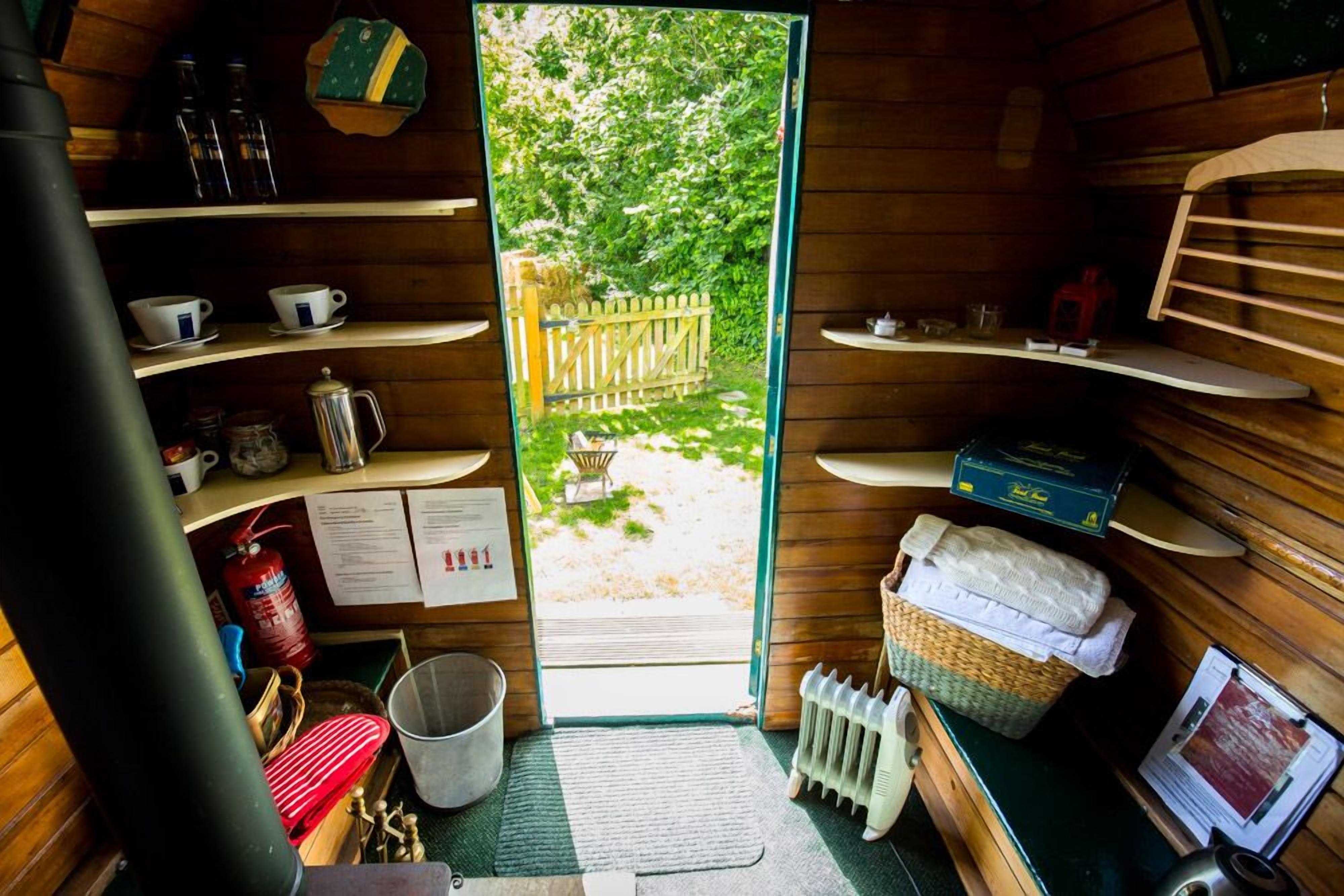 Il s’agit d’une petite pièce de style cabane, avec des murs en bois et une porte donnant sur un espace extérieur. Elle comprend une étagère avec des tasses et une bouilloire, un panier avec des serviettes et un radiateur.