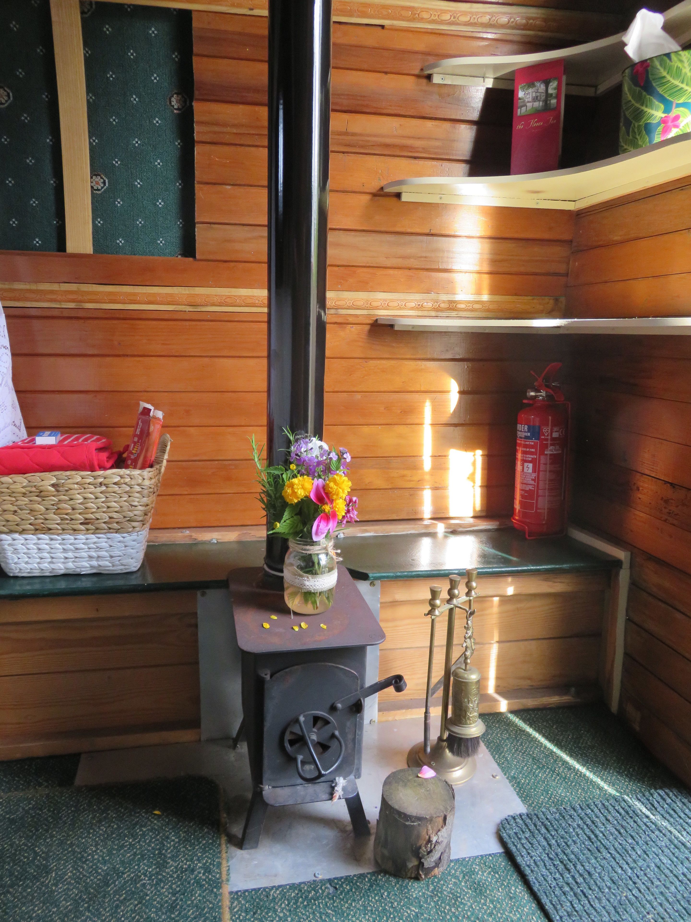 Interior de madera con estufa de leña, una cesta con objetos, un florero con flores y un extintor.