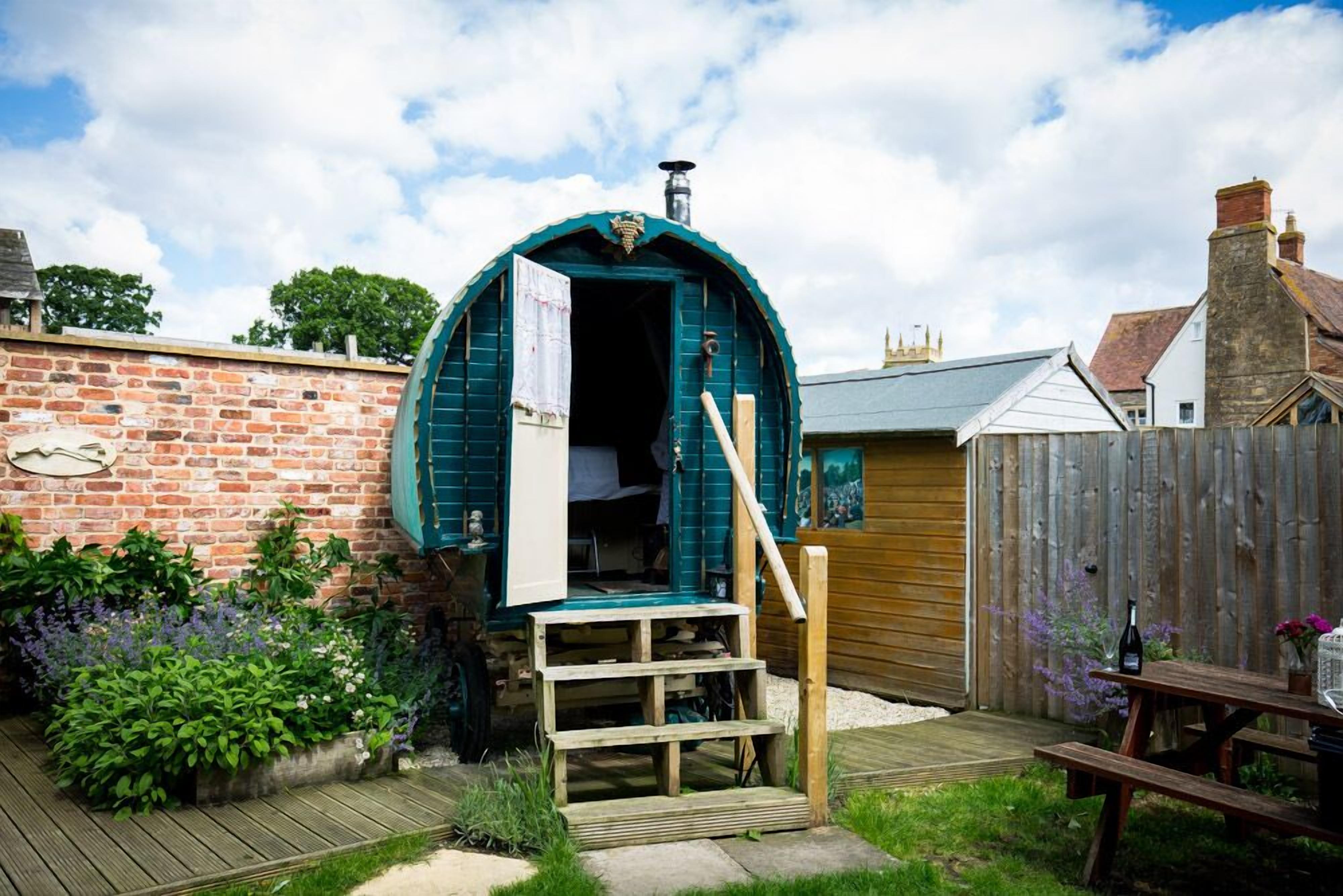 A rustic, green, dome-shaped structure with a wooden staircase leading to an open door, set in a backyard with a brick wall, wooden fence, and a picnic table.