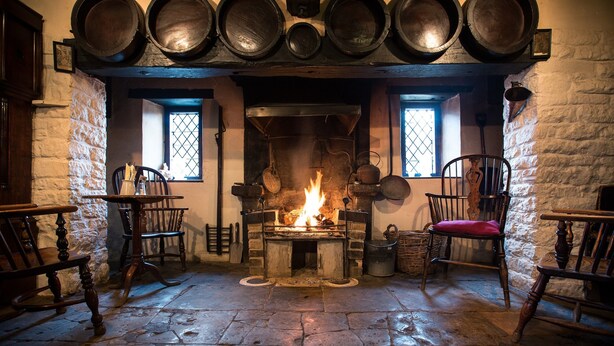 Una habitación con suelo de piedra, chimenea, sillas de madera y una mesa.