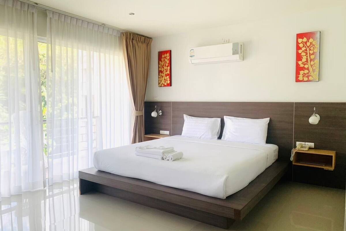 A modern hotel room with a large bed, a bedside table, a nightstand, a wall-mounted air conditioner, and a window with sheer curtains.