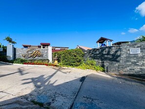 Uma entrada com portão e muro de pedra, um letreiro com a inscrição "Assembly Point" e um edifício com telhado vermelho ao fundo.