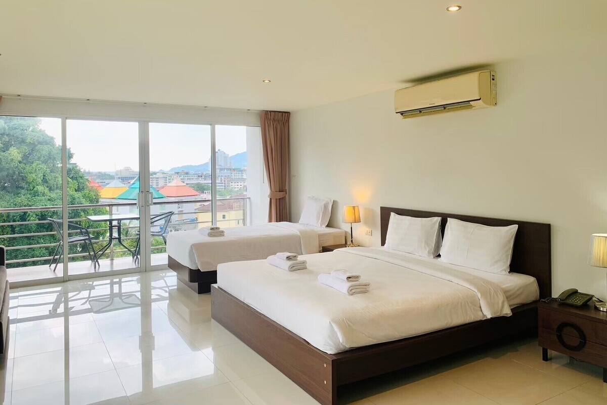 A hotel room with a large bed, a smaller bed, a balcony with a table and chairs, and a city view.