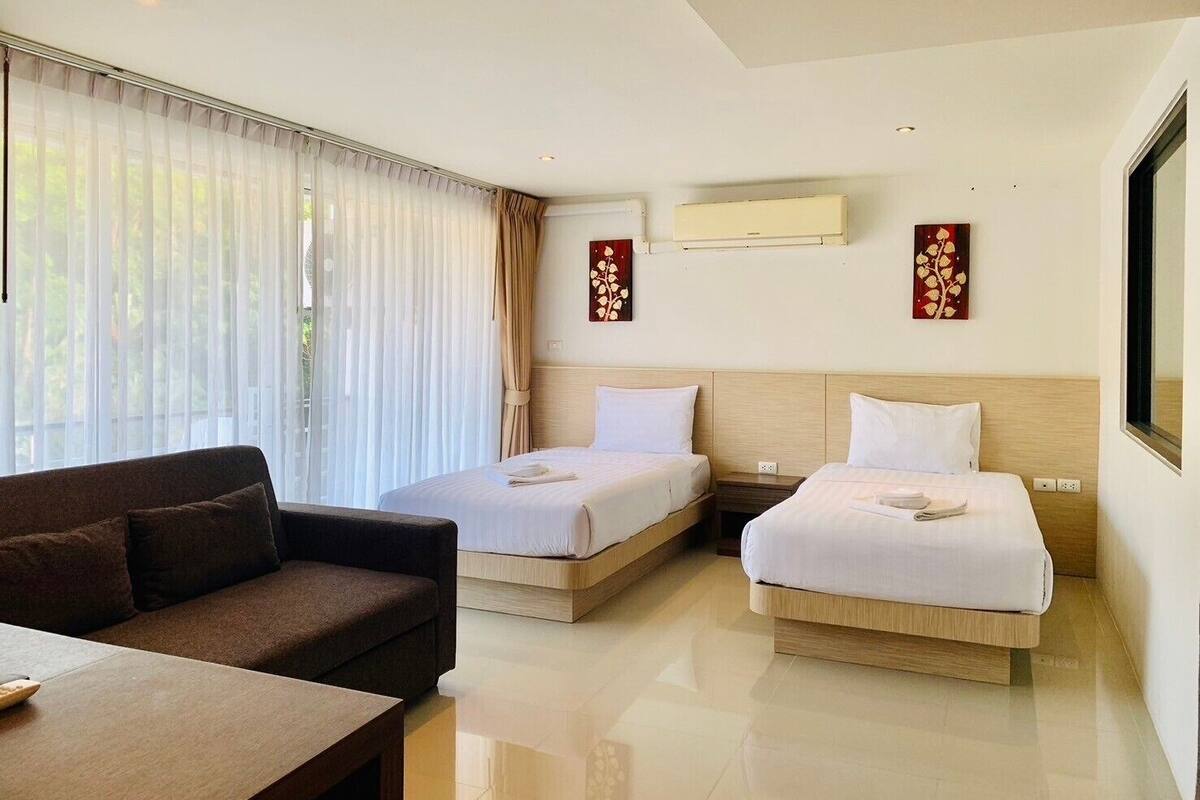 A hotel room with two single beds, a sofa, a table, and a large window with sheer curtains.