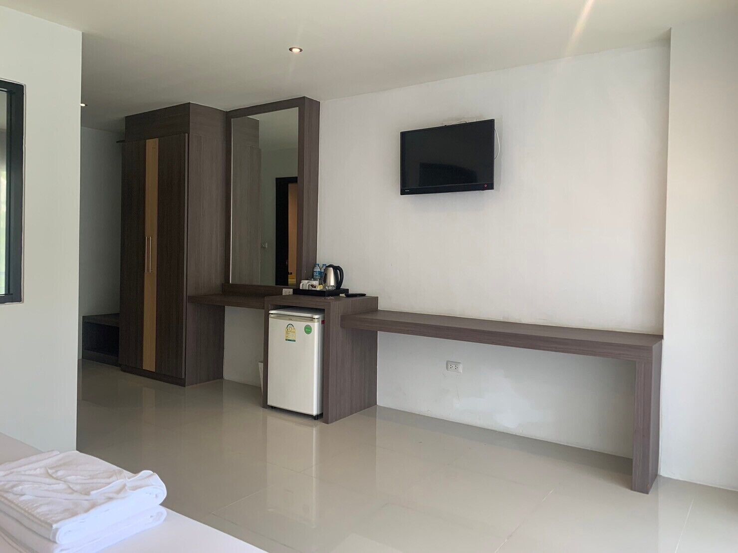 A modern hotel room with a large mirror, a flat-screen TV mounted on the wall, a small refrigerator, and a neatly made bed.
