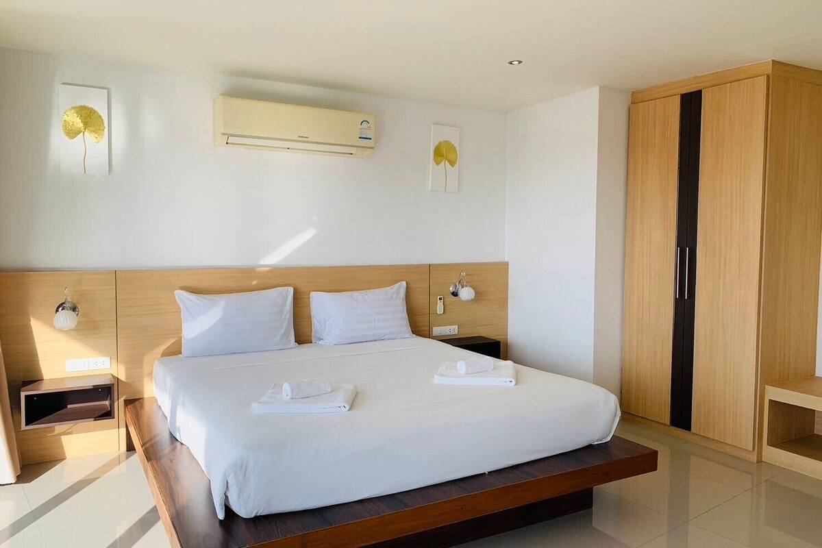 A modern hotel room with a large bed, wooden headboard, bedside tables, an air conditioning unit, and a wardrobe.