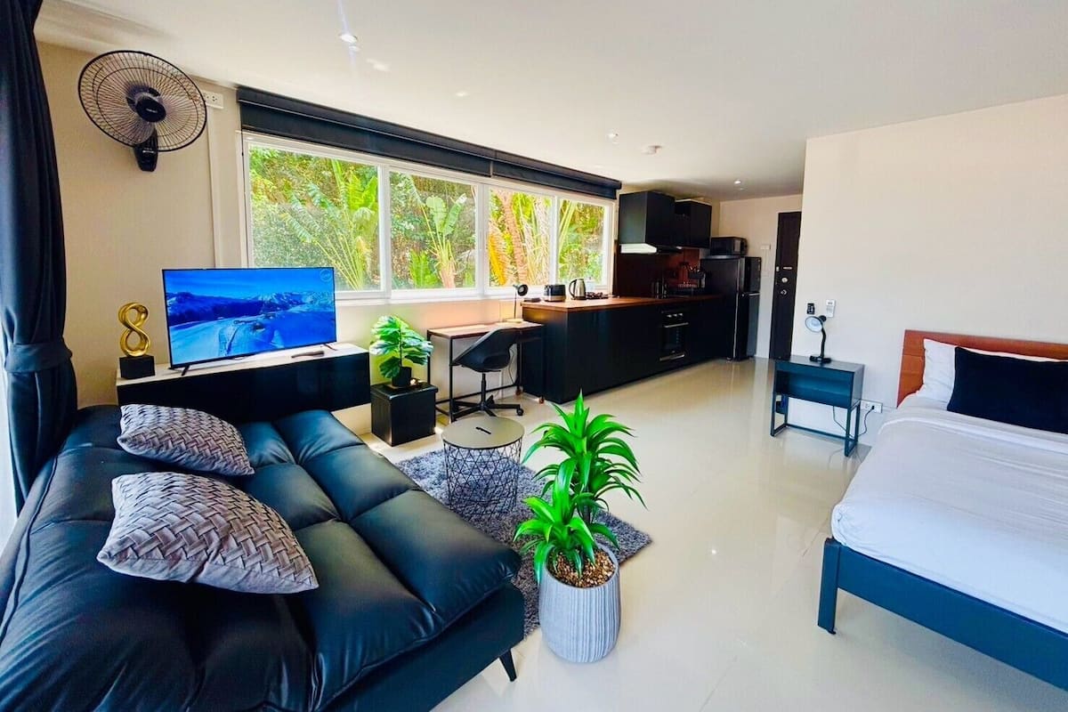 A modern hotel room with a large bed, a sofa, a desk, a TV, and a view of greenery through the windows.