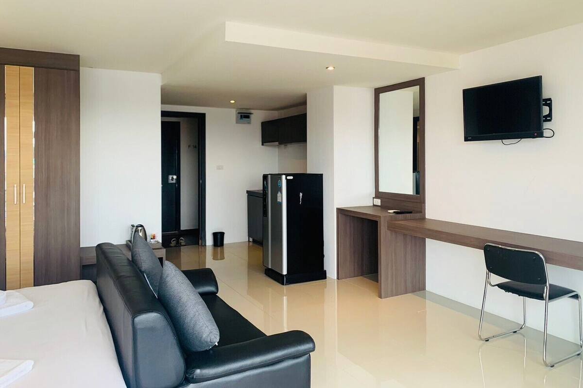 A modern hotel room with a black leather sofa, a flat-screen TV mounted on the wall, a kitchen area with a refrigerator, and a wooden cabinet.